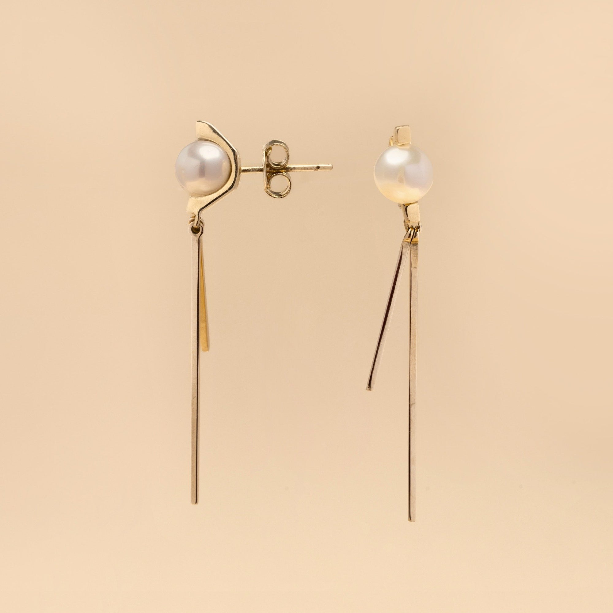 Tick & Tack Örhängen i 14K Guld – pärla med halvcirkel och två hängande linjer, flatlay mot beige bakgrund.